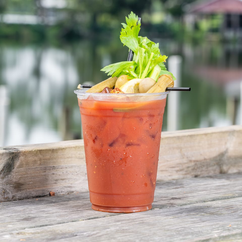Bloody Mary cocktail with celery, lemon, okra, and mushrooms on a wooden dock by water.