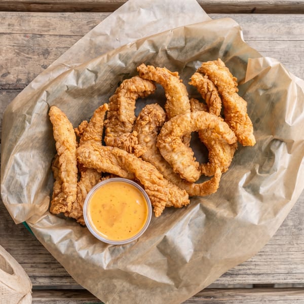 Crispy fried chicken tenders and a dipping sauce in a paper-lined basket on a wooden table.