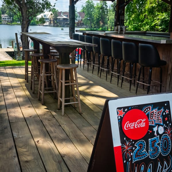 Outdoor bar with stools and tables on a wooden deck overlooking a lake with houses.