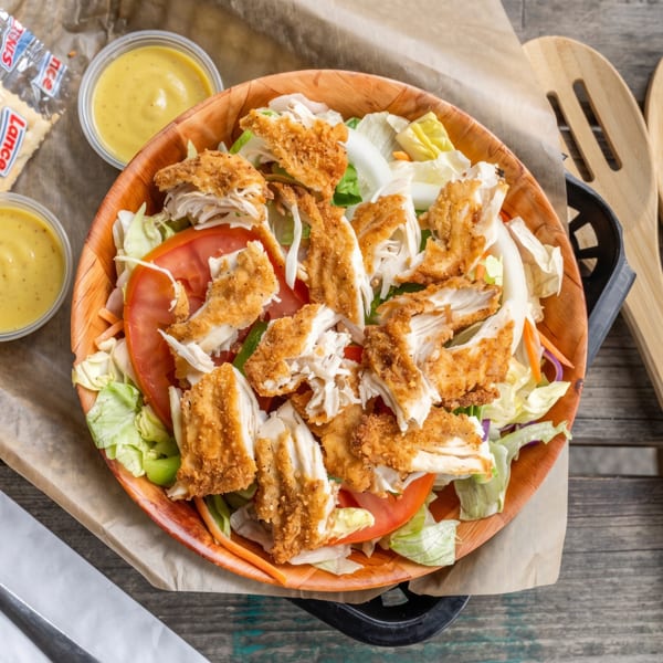 Crispy chicken salad with lettuce, tomatoes, and dressing, served with crackers and wooden utensils.