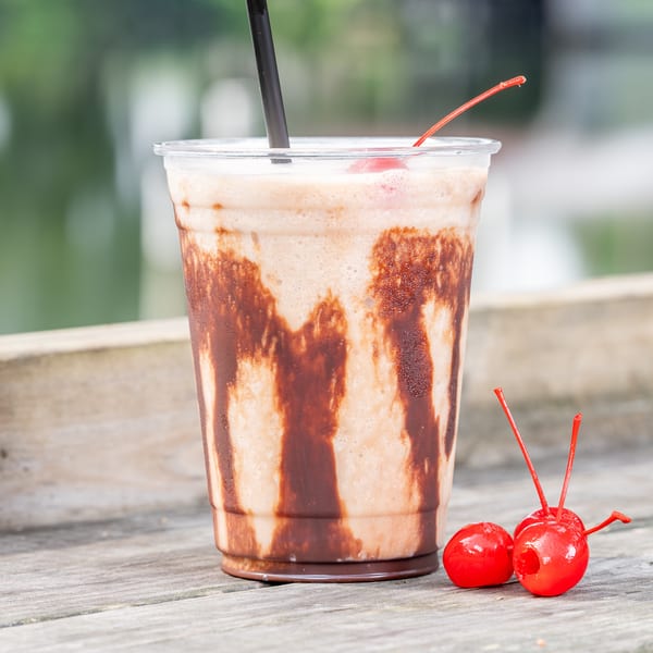 Chocolate milkshake with chocolate syrup streaks, a cherry, and a straw on a wooden surface.